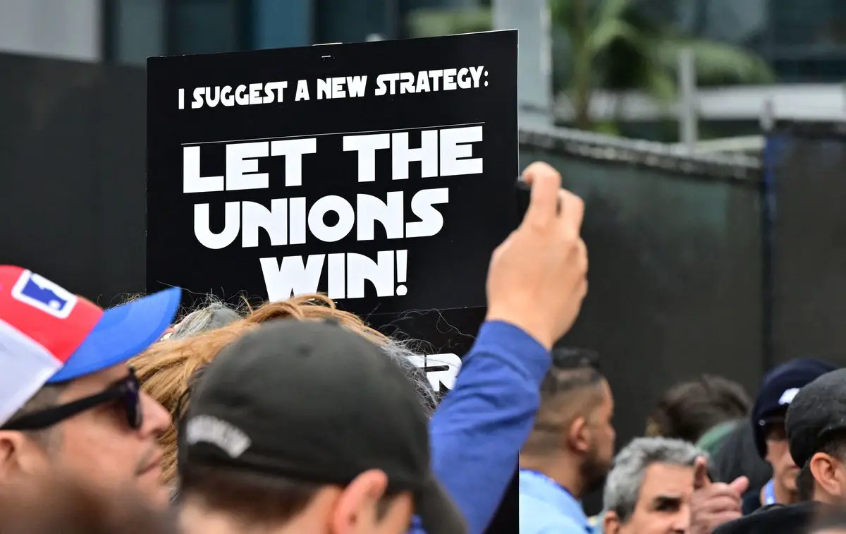 Los manifestantes sostienen carteles mientras hacen piquetes durante la huelga continua del Writers Guild of America (WGA) en Los Ángeles, California, el 26 de mayo de 2023. En el centro de la imagen hay un cartel en la fuente del título de Star Wars que dice 